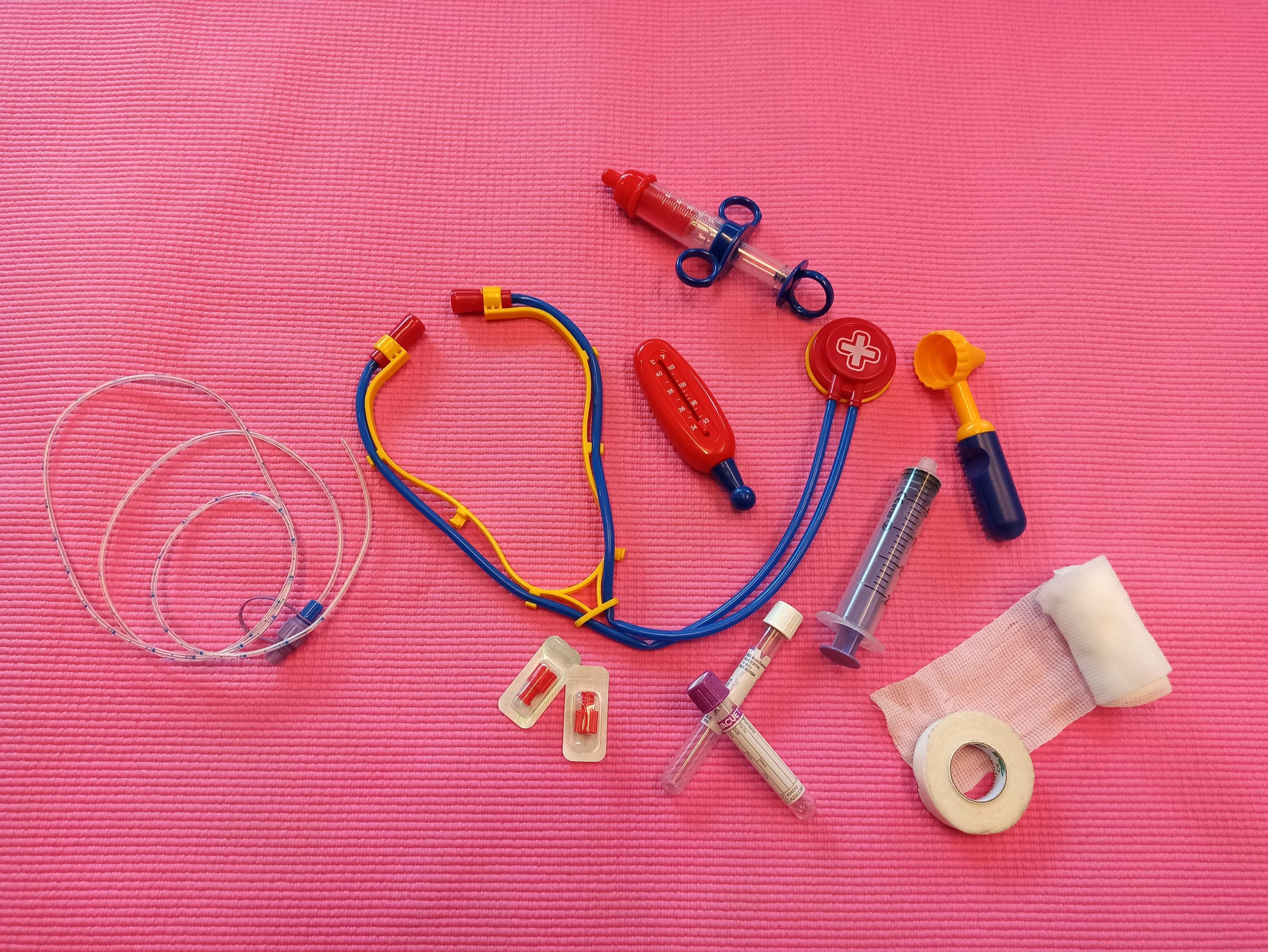 Stetoskooppi, lääkeruiskuja, sidetarpeita ja muita sairaalavälineitä vaaleanpunaisella alustalla.